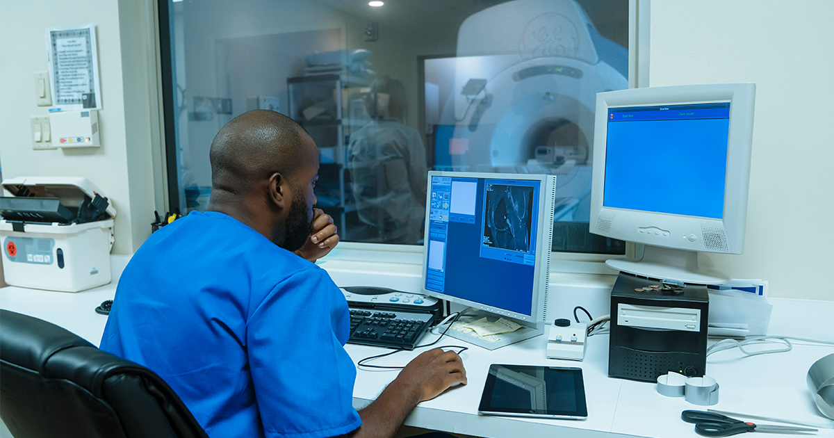 Healthcare worker looking at computer screen with MRI room in background