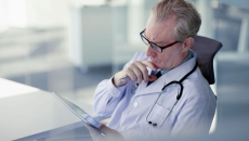 Male doctor with glasses sits at a desk looking at a tablet