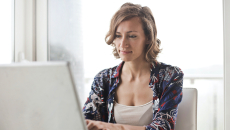 Woman sits at a table and is reading and typing on a laptop