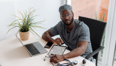 Doctor in scrubs sits at a desk near a patio with stethescope, tablet, laptop and phone.