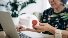A patient holding a bottle of medication faces a laptop computer in an online consultation