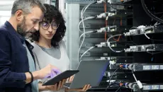 IT staff in a server room look at data on a laptop and tablet 