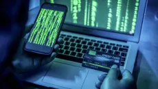 Person holding a smartphone and credit card in front of laptop screen in dark room