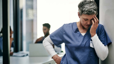 Nurse with short grey hair leans against a wall rubbing her temple