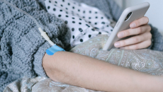 Patient in a blue sweater comforter recuperates at home and receives an IV infusion while checking patient portal information on a cell phone.