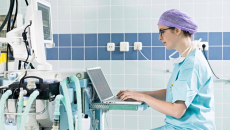 Healthcare worker in scrubs using laptop next to healthcare machinery