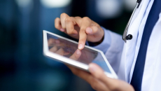 Close up of a doctor, wearing a lab coat and stethoscope, accesses healthcare IT from a tablet.