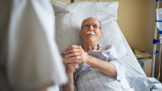 A patient in a hospital bed smiles and clasps the hand of a clinician.