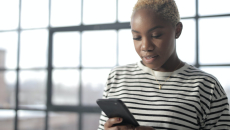 A woman of color with a striped shirt looks happily at her mobile phone.