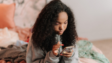 Young woman wearing a sweatshirt is sitting on her bed and checking a mobile application for information.