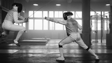 Black and white image of two individuals fencing. One is jumping but the opponent finds the weak spot.