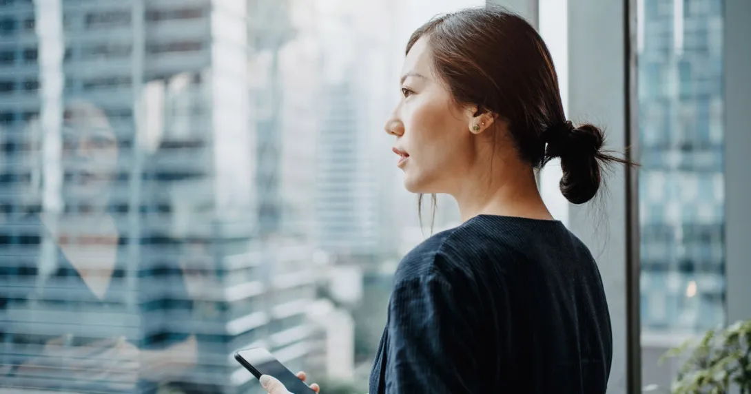 A woman holding a smartphone looks outside a window