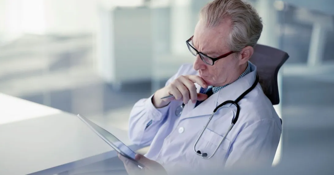 Male doctor with glasses sits at a desk looking at a tablet