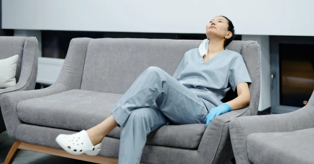 Female nurse in grey scrubs on rests on a couch with her head back