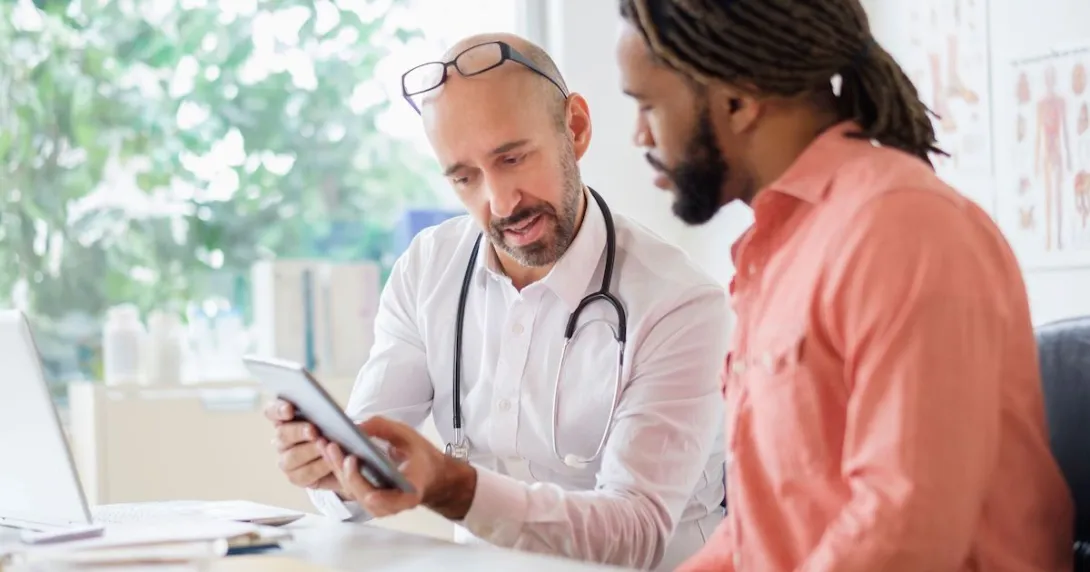 A male doctor sits with another man and they are looking at SDOH data on a mobile device.