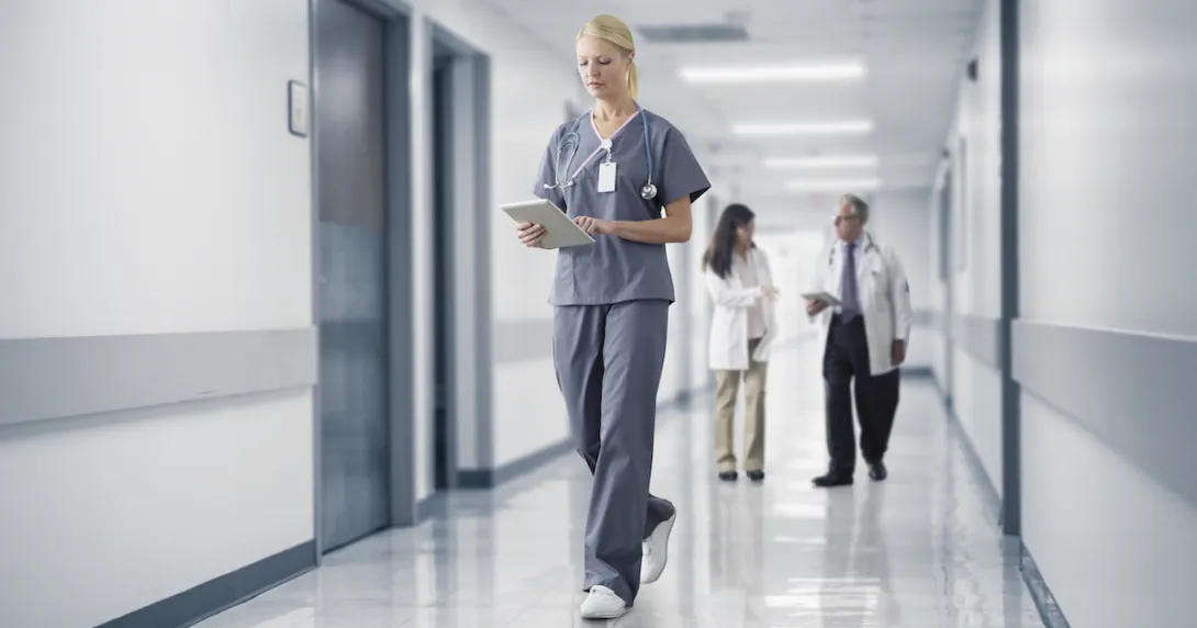 Nurses and doctors walk down a hospital hallway, one checks patient monitoring data on a tablet.