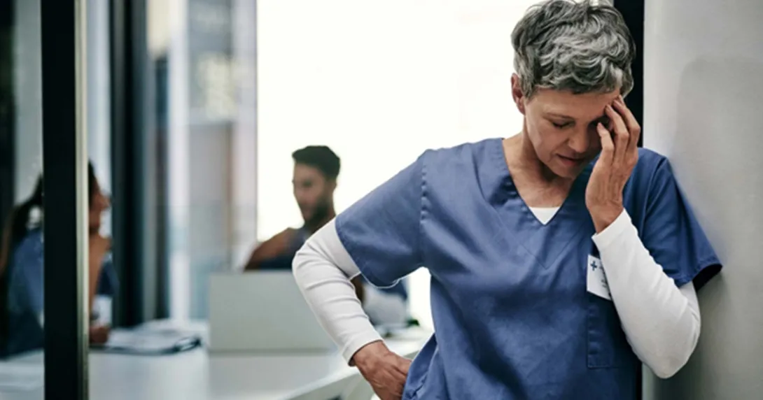 Nurse with short grey hair leans against a wall rubbing her temple