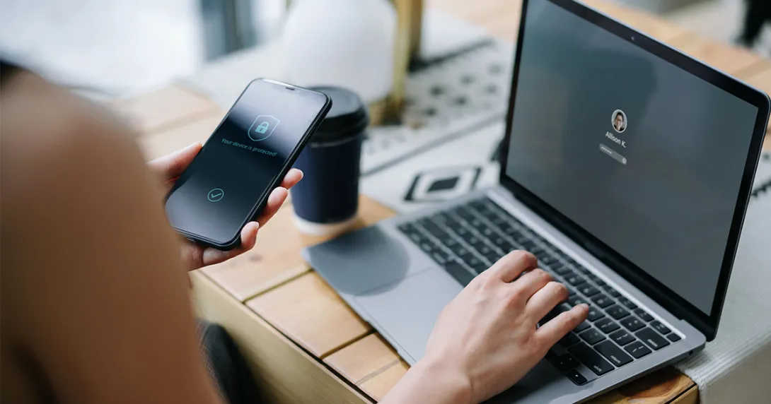An individual is holding her cell phone and logging into her laptop.