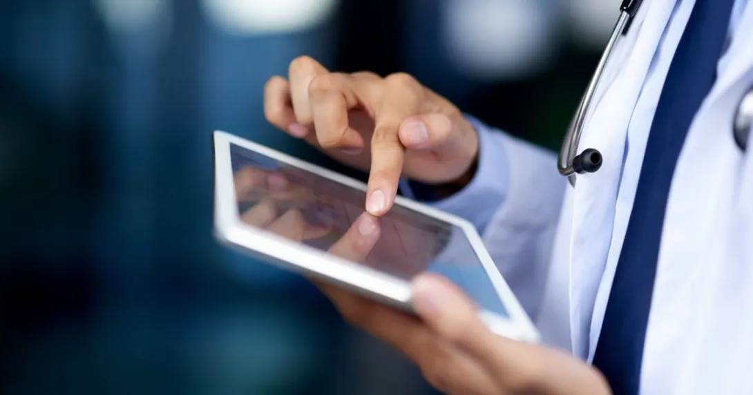 Close up of a doctor, wearing a lab coat and stethoscope, accesses healthcare IT from a tablet.