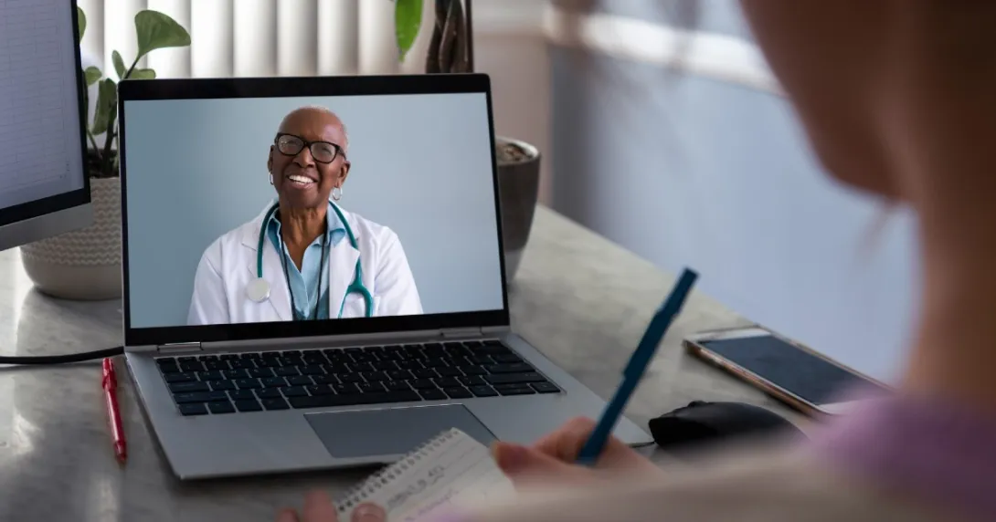 Female doctor on laptop smiles at patient in foreground who is taking notes