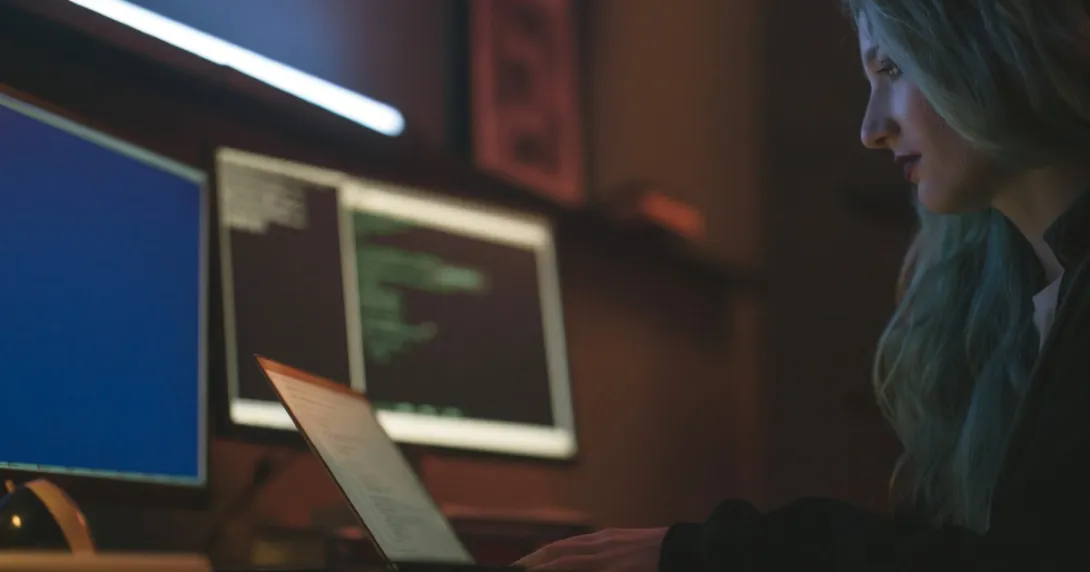 Person with long hair sits before a laptop and multiple screens