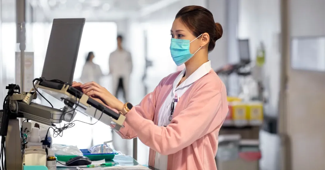 Nurse in mask and pink sweater types notes in a hospital hallway