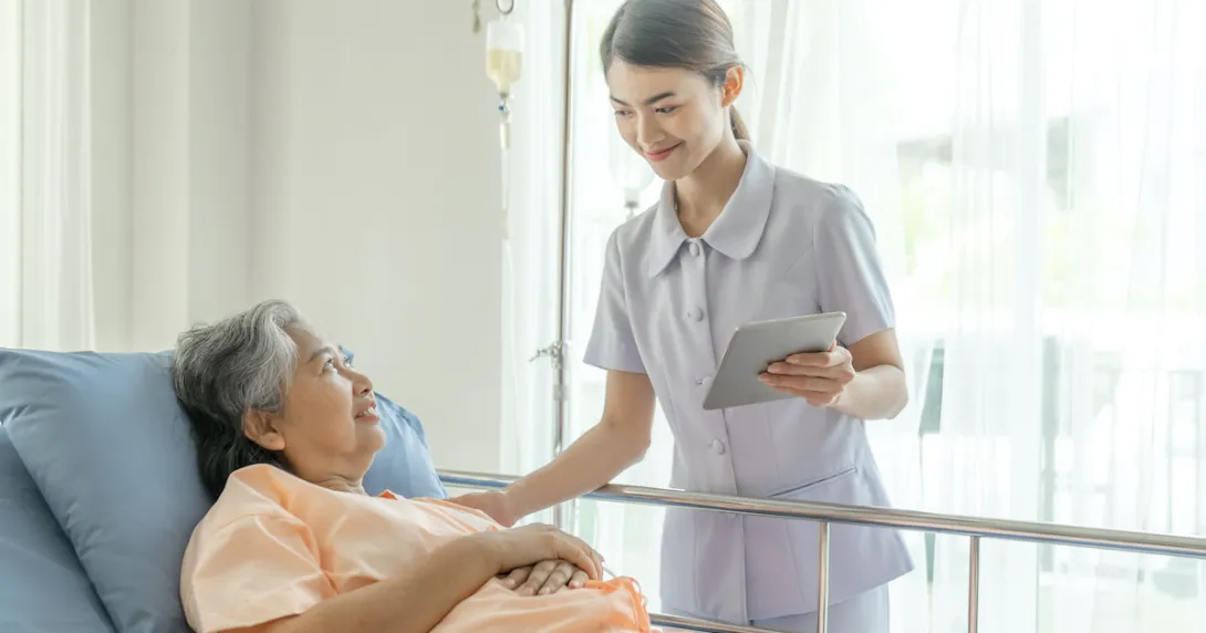 A nurse holding a digital tablet is assisting a patient lying in bed
