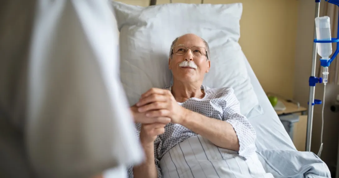 A patient in a hospital bed smiles and clasps the hand of a clinician.