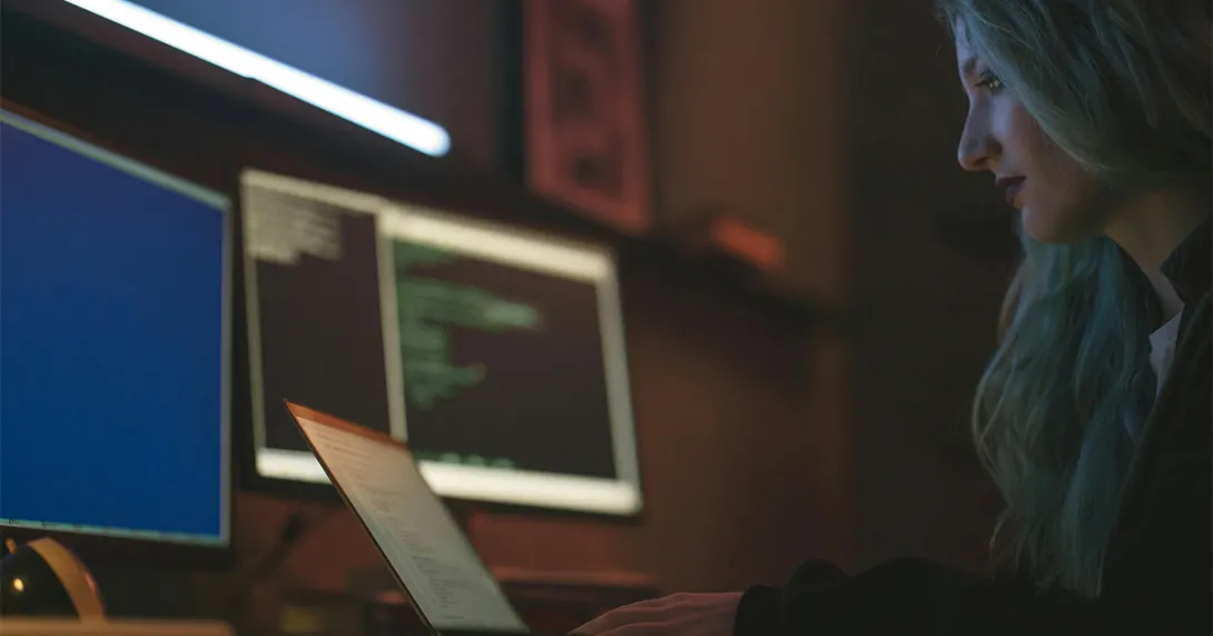 A security IT professional with long hair sits at a desk and monitors a client system remotely
