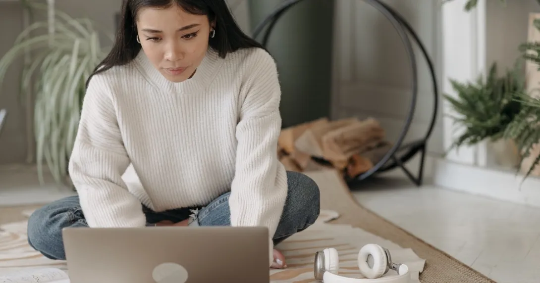 A woman with long hair sits on her floor at home as she engages in a behavioral health appointment.